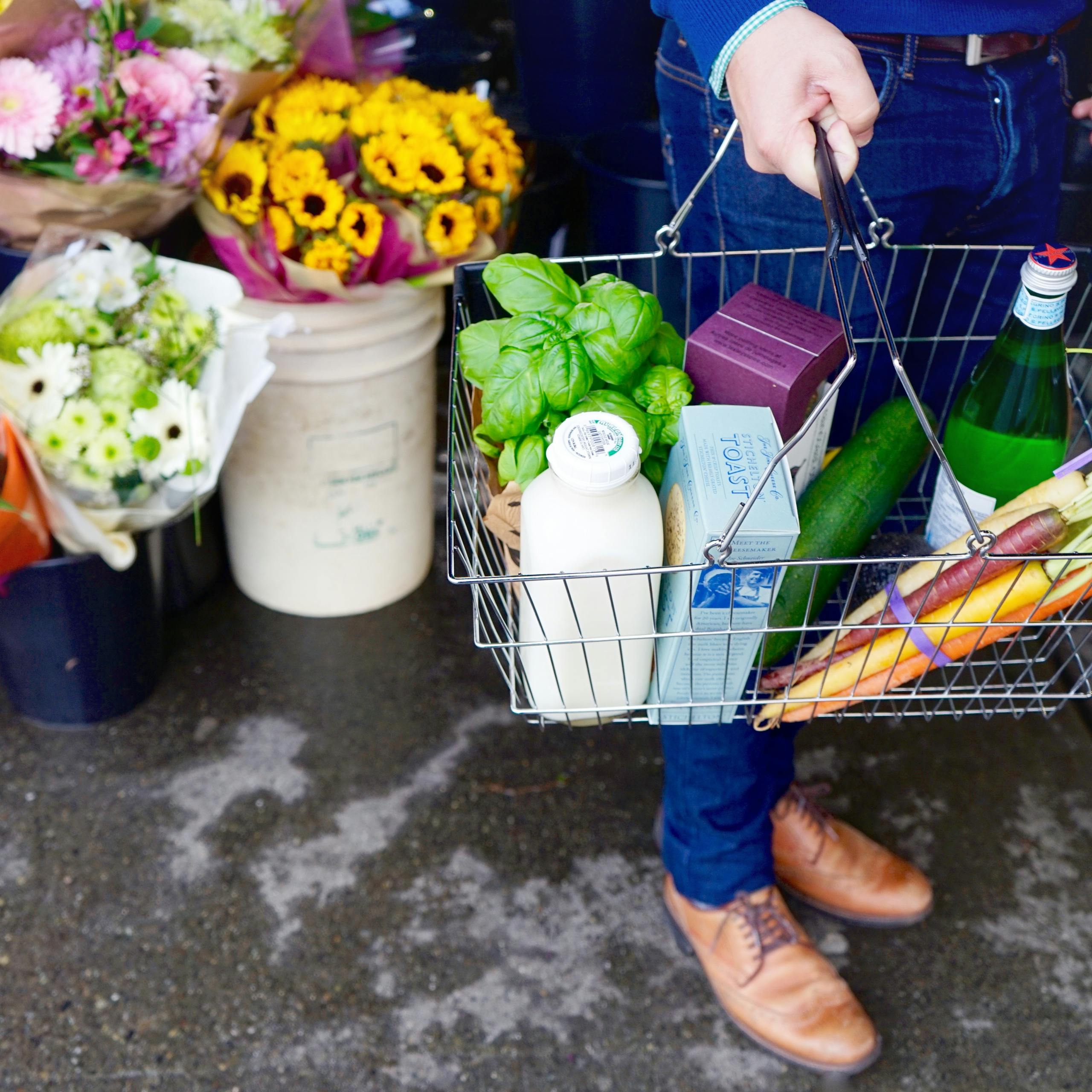 Close-up of a grocery basket filled with fresh produce, milk, and flowers in a market setting.